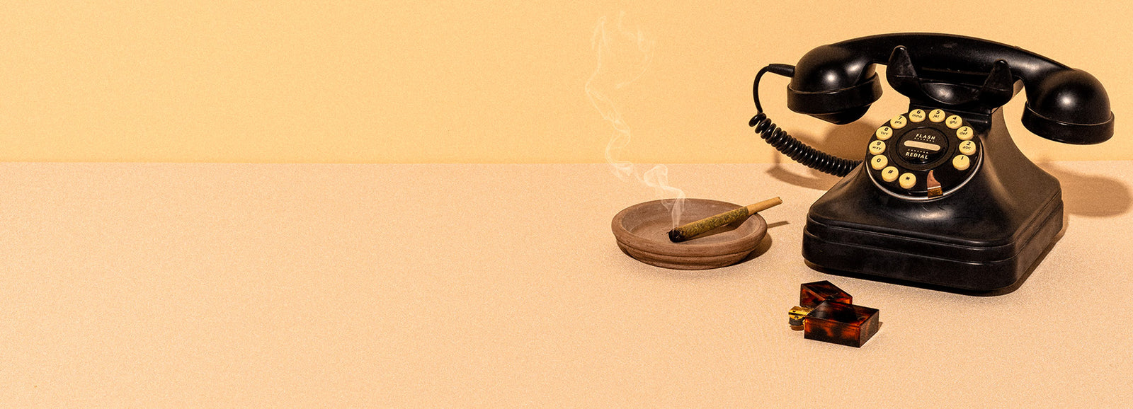 An old school rotary phone and lit joint rest on a tabletop.